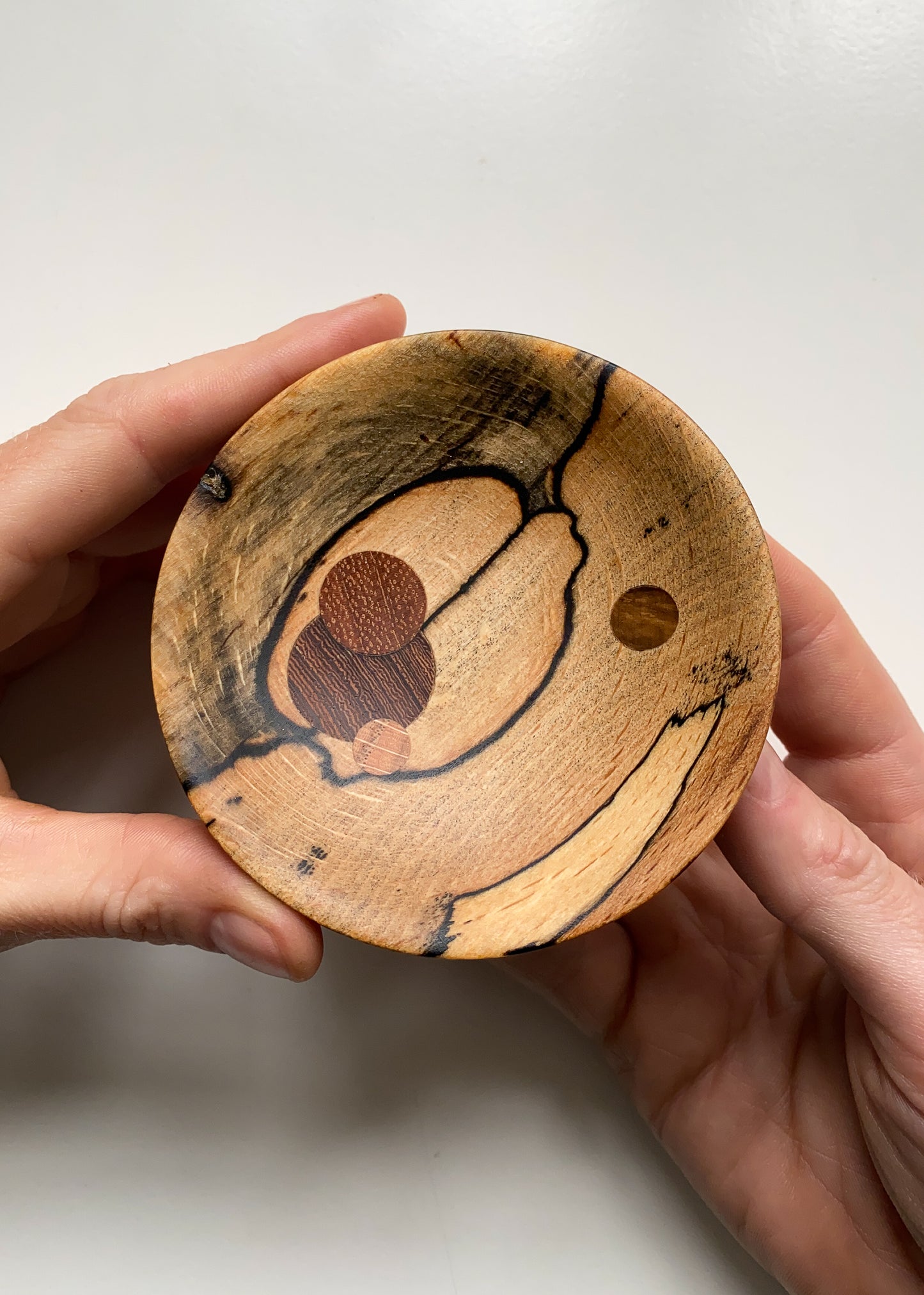 Small spalted wooden bowl with circular wooden inlays held in hands against white background.