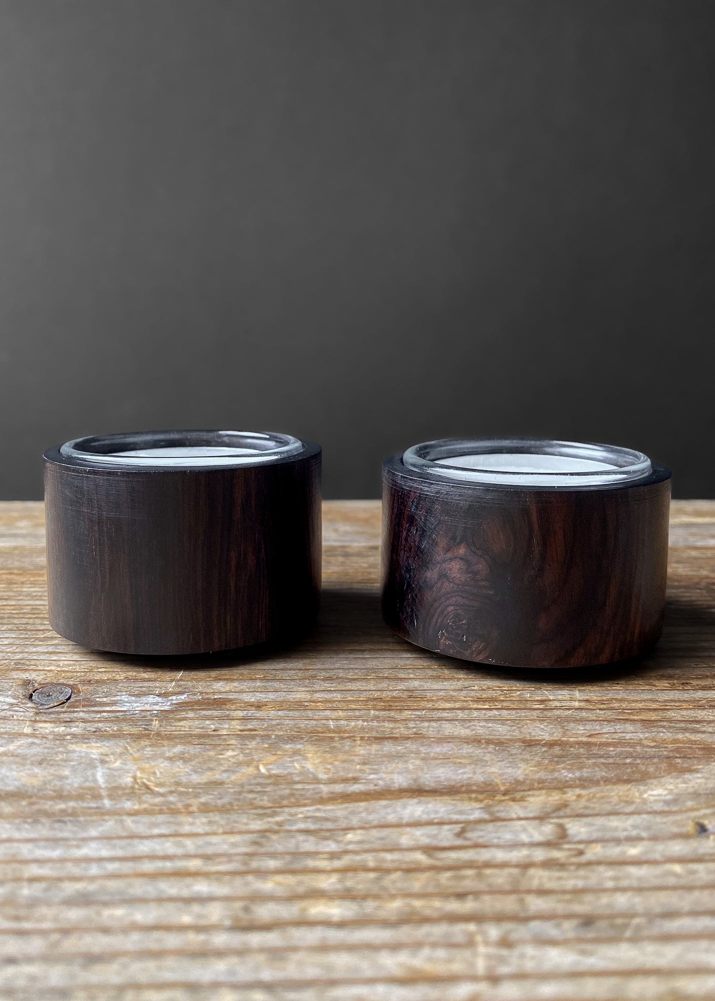 Pair of modern, round wooden candle holders with glass inserts, displayed on a rustic wooden table against a dark background.