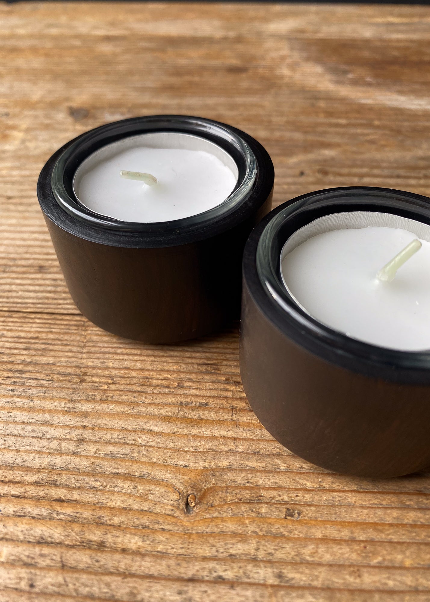 Close-up view of two dark wood and glass tealight candle holders, each holding a white candle with a visible wick, placed on a textured wooden surface.