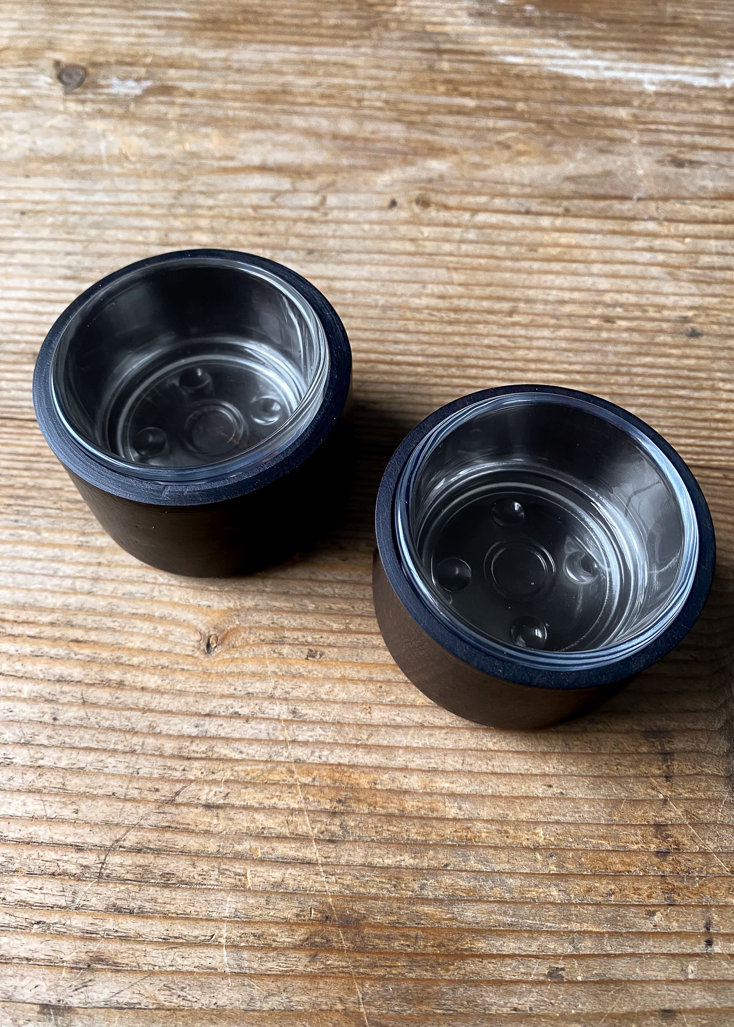 Overhead view of two empty wooden candle holders with clear glass inserts, showing the circular glass bases and the sleek, cylindrical wood exteriors on a wooden tabletop.