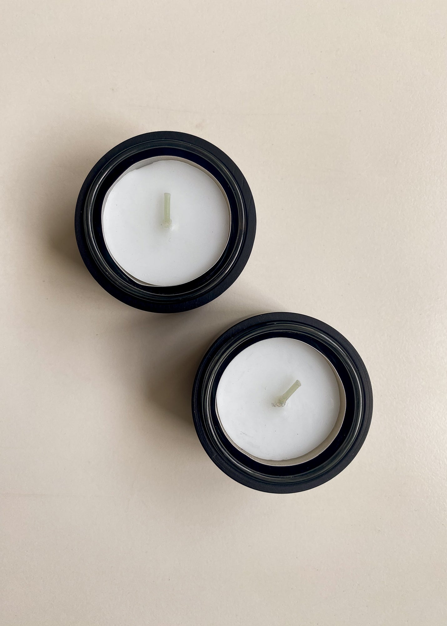 Minimalist top-down view of two white tealight candles placed inside round, black wooden holders on a light cream background.