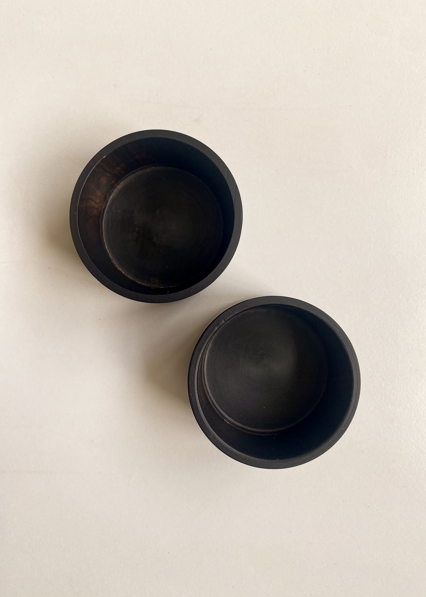 Overhead shot of two empty, round black wooden candle holders with smooth interiors, arranged on a neutral light surface.