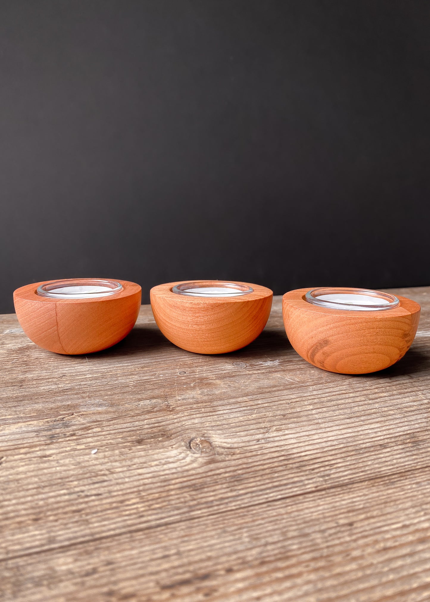 Set of three rounded cherry wood candle holders with glass inserts and tea light candles on a dark background.