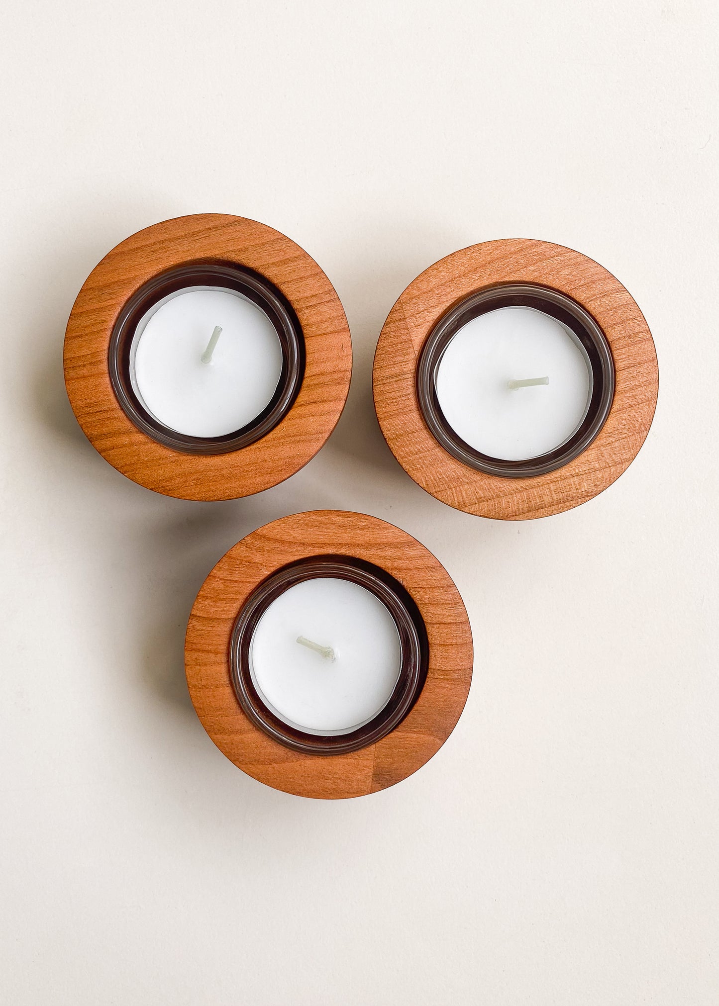 Overhead view of three rounded cherry wood candle holders with glass inserts and tea light candles on a white background.