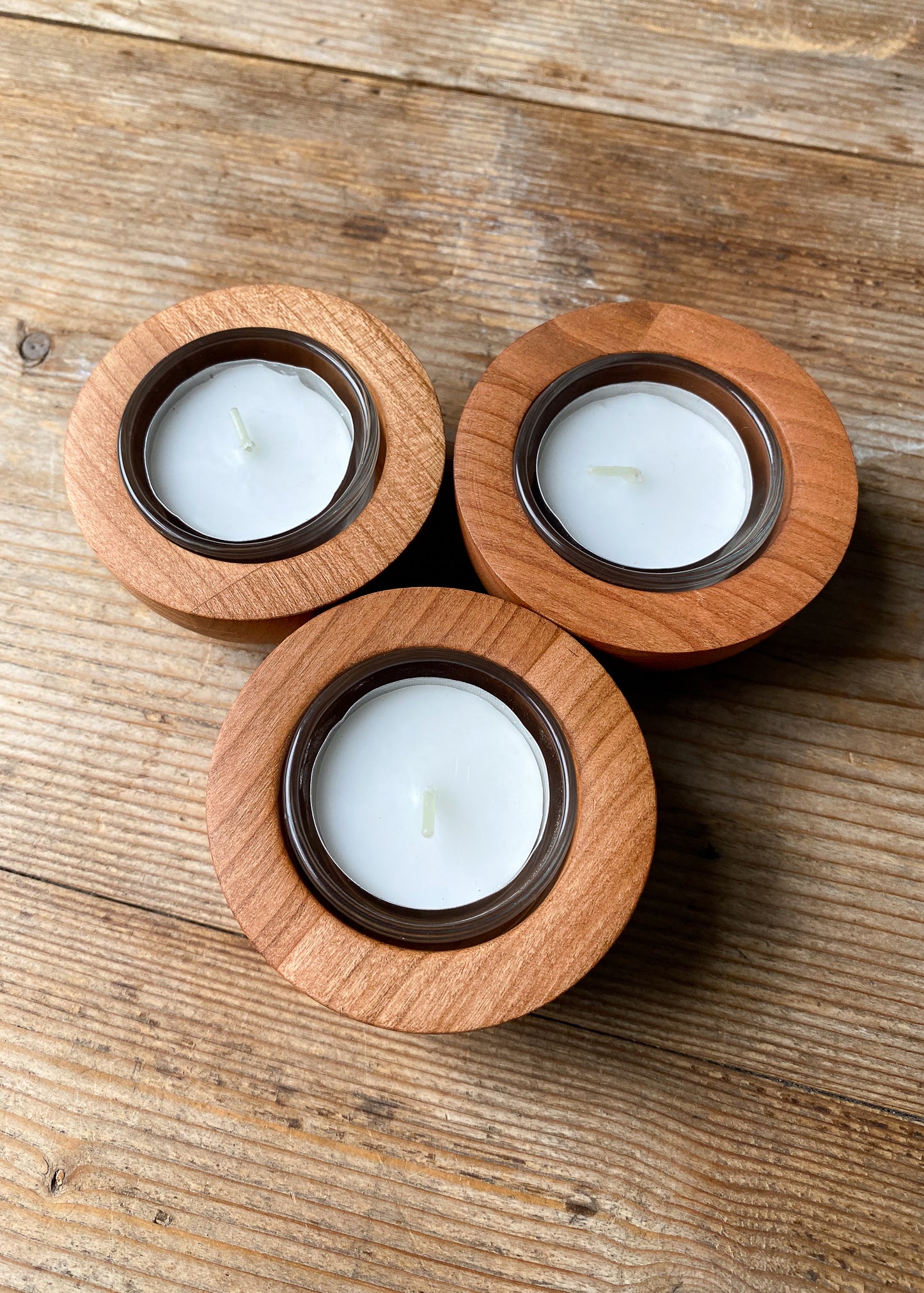 Three rounded cherry wood candle holders with glass inserts and tea light candles on a wooden table.
