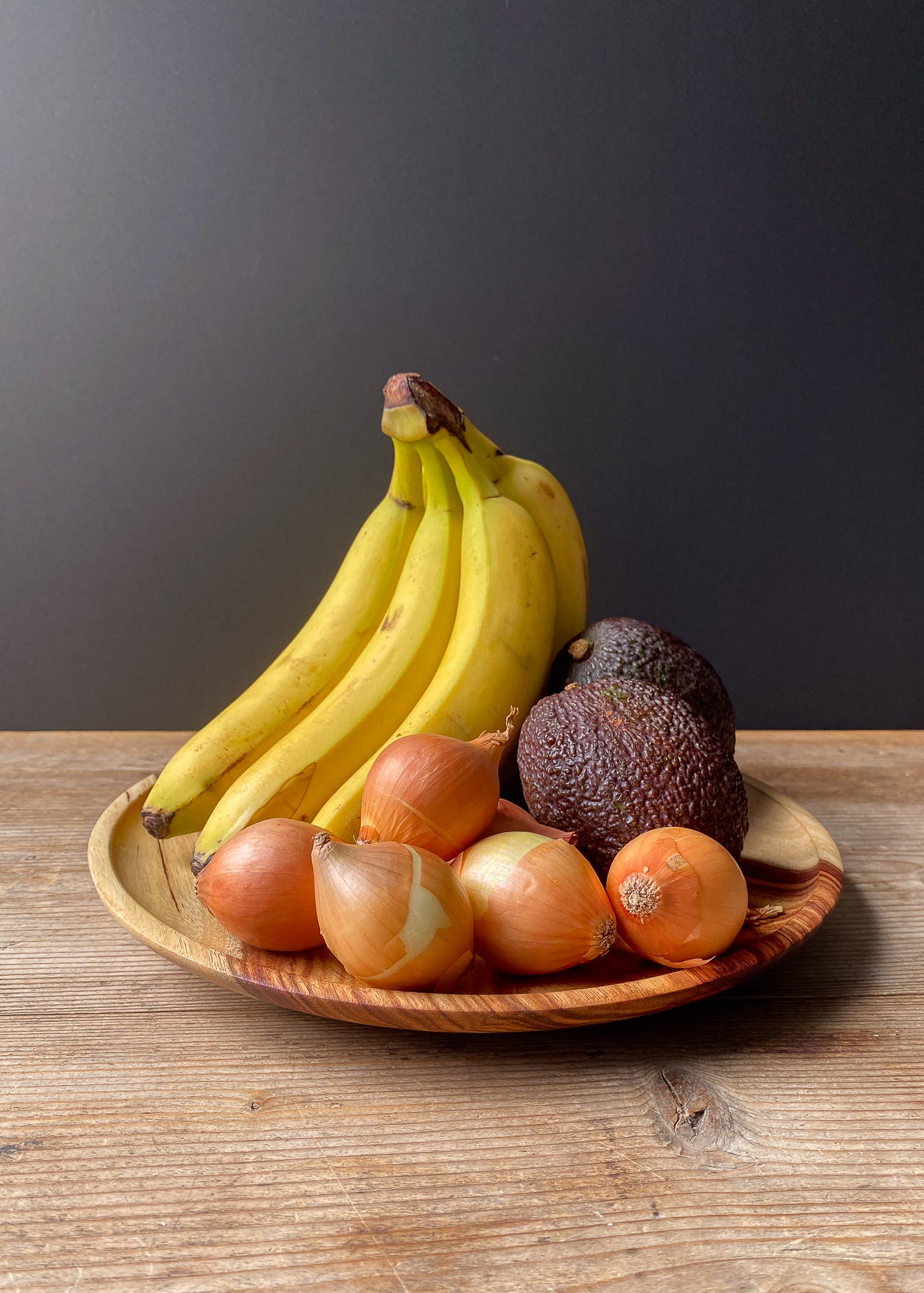 Bananas, avocados, and onions on a wooden tray with a dark background.