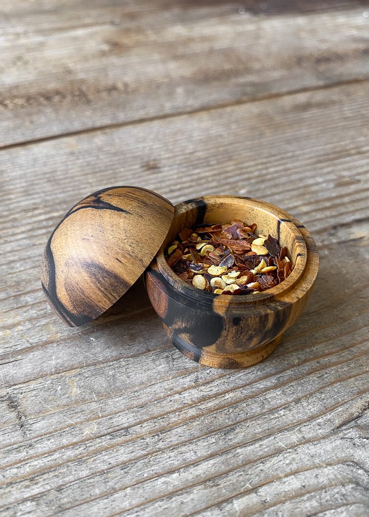 Small orange and black wood lidded box on wooden table with open lid displaying chili flakes.