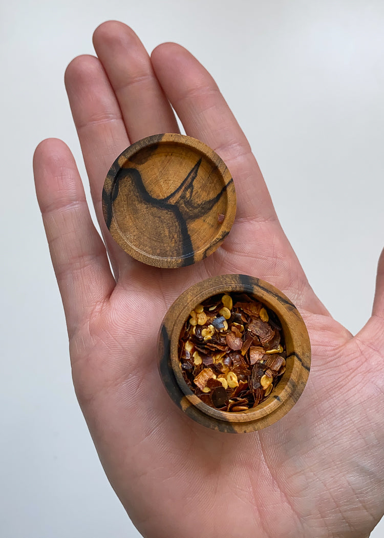 Overhead view of patterned wooden lidded box held open in hand displaying chili flakes against white background.