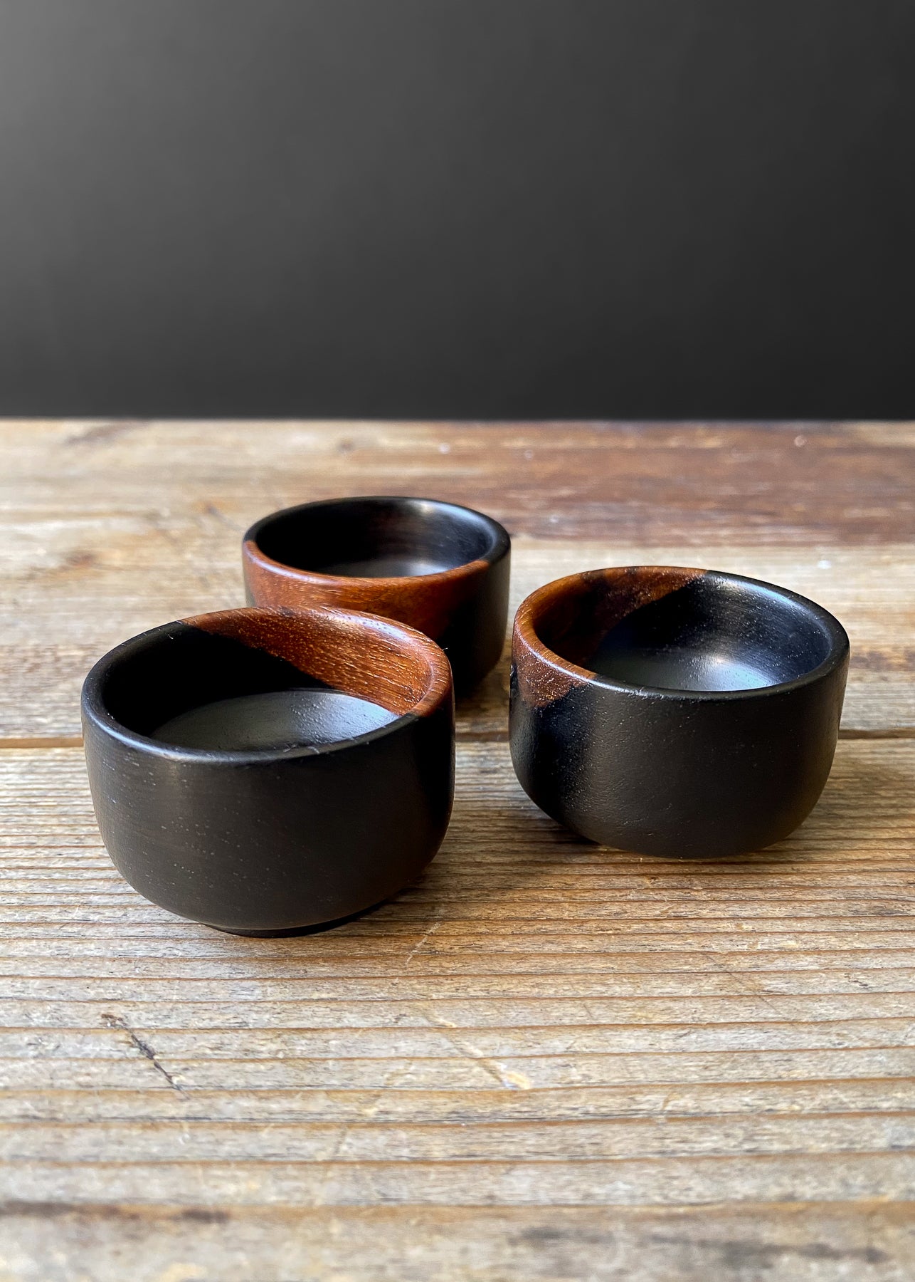 Three small black wooden bowls on wood 
table with black background.