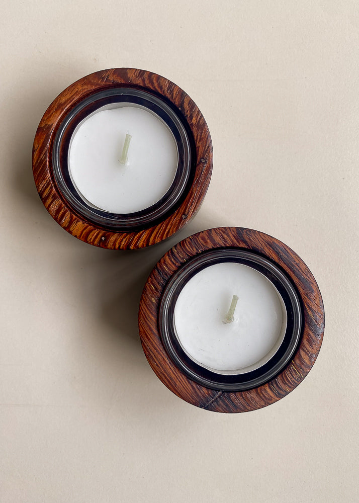 Overhead view of two textured wooden candle holders with glass inserts and tea light candles on white background.
