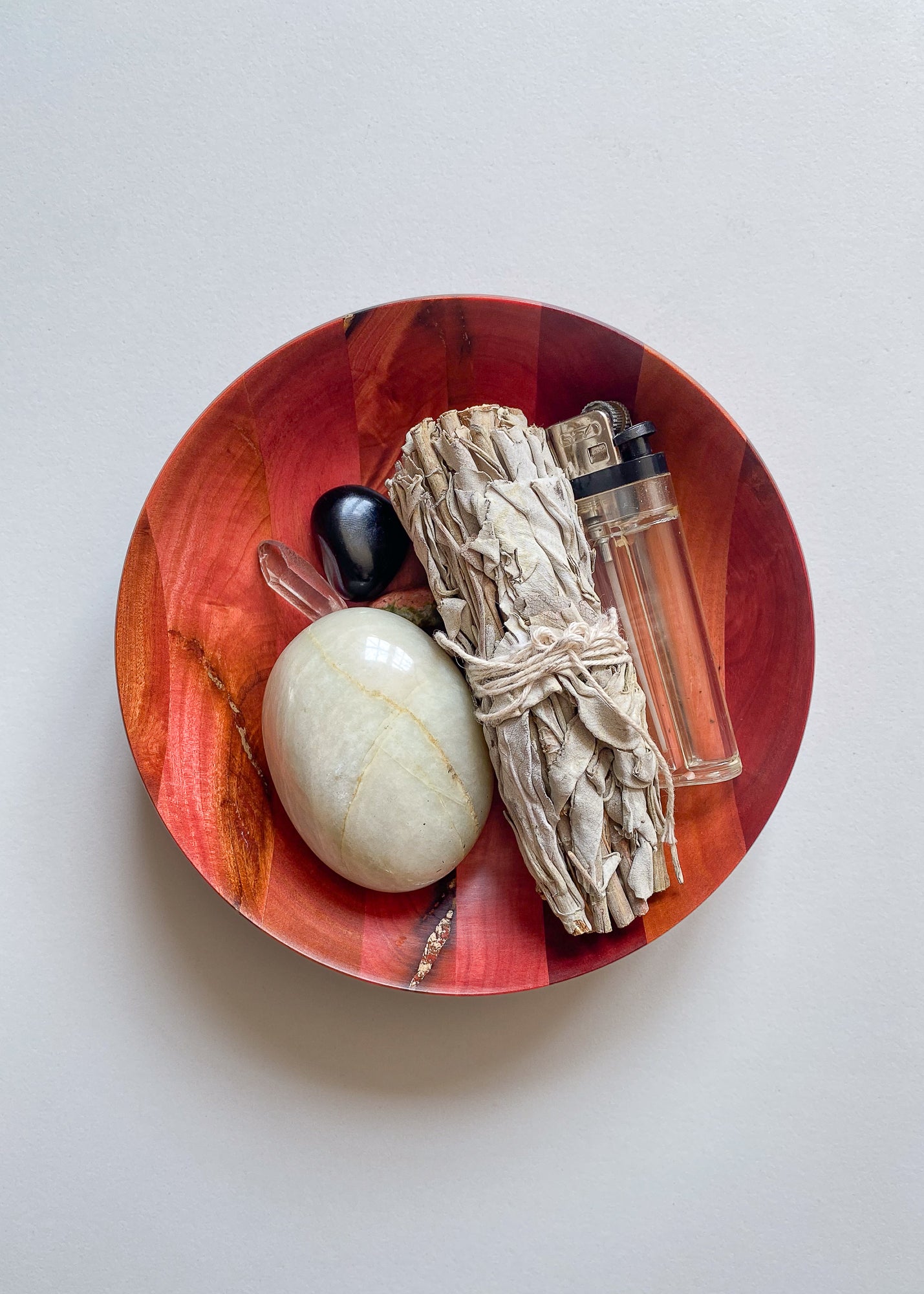 Overhead view of pink wooden bowl with sage bundle and crystals on white background.