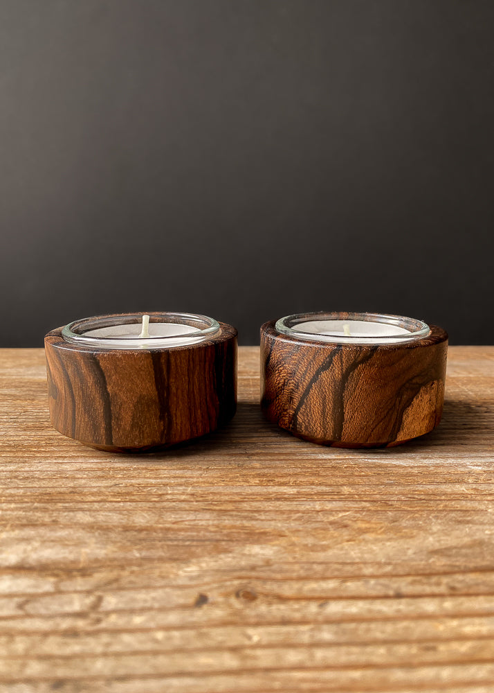 Two Ziricote candle holders with glass inserts and tea light candles on wooden table.