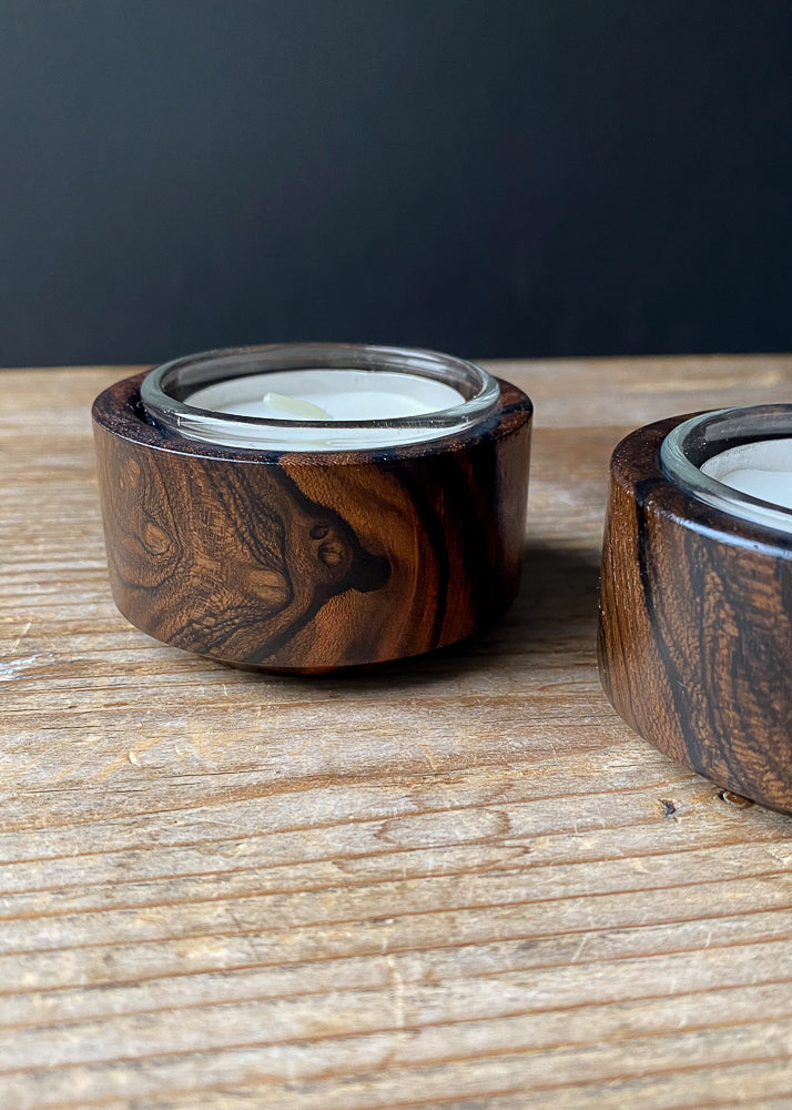 Close up view of two Ziricote candle holders with glass inserts and tea light candles on a wooden table.