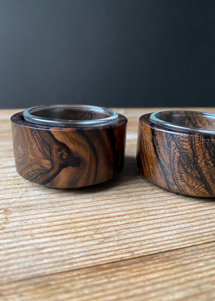 Close up side view of two Ziricote candle holders with glass inserts on a wooden table.