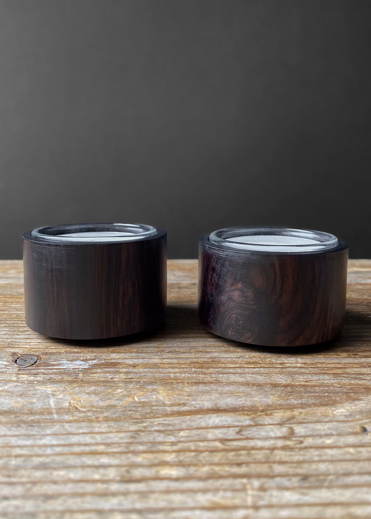 Pair of modern, round wooden candle holders with glass inserts, displayed on a rustic wooden table against a dark background.