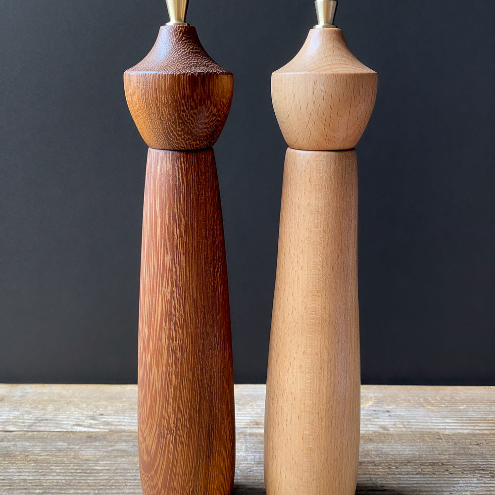 Two wooden pepper mills, one brown and one beige, on a wooden surface with a dark background.