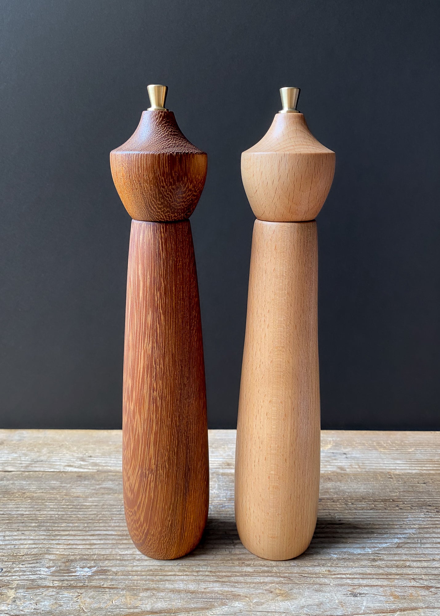 Two wooden pepper mills, one brown and one beige, on a wooden surface with a dark background.