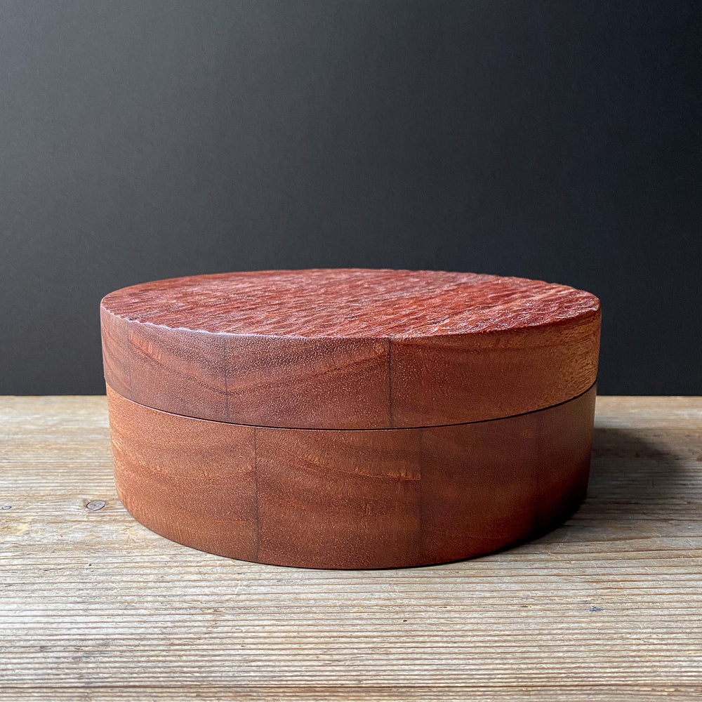 Large round wooden box with textured lid on a wooden surface with a dark gray background.