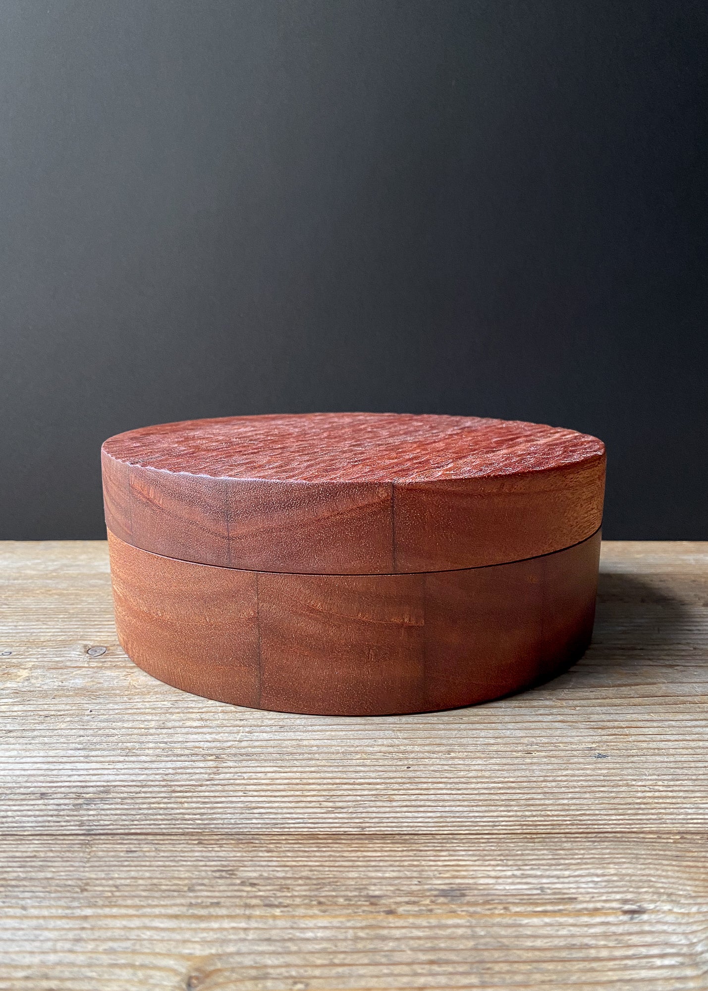 Large round wooden box with textured lid on a wooden surface with a dark gray background.