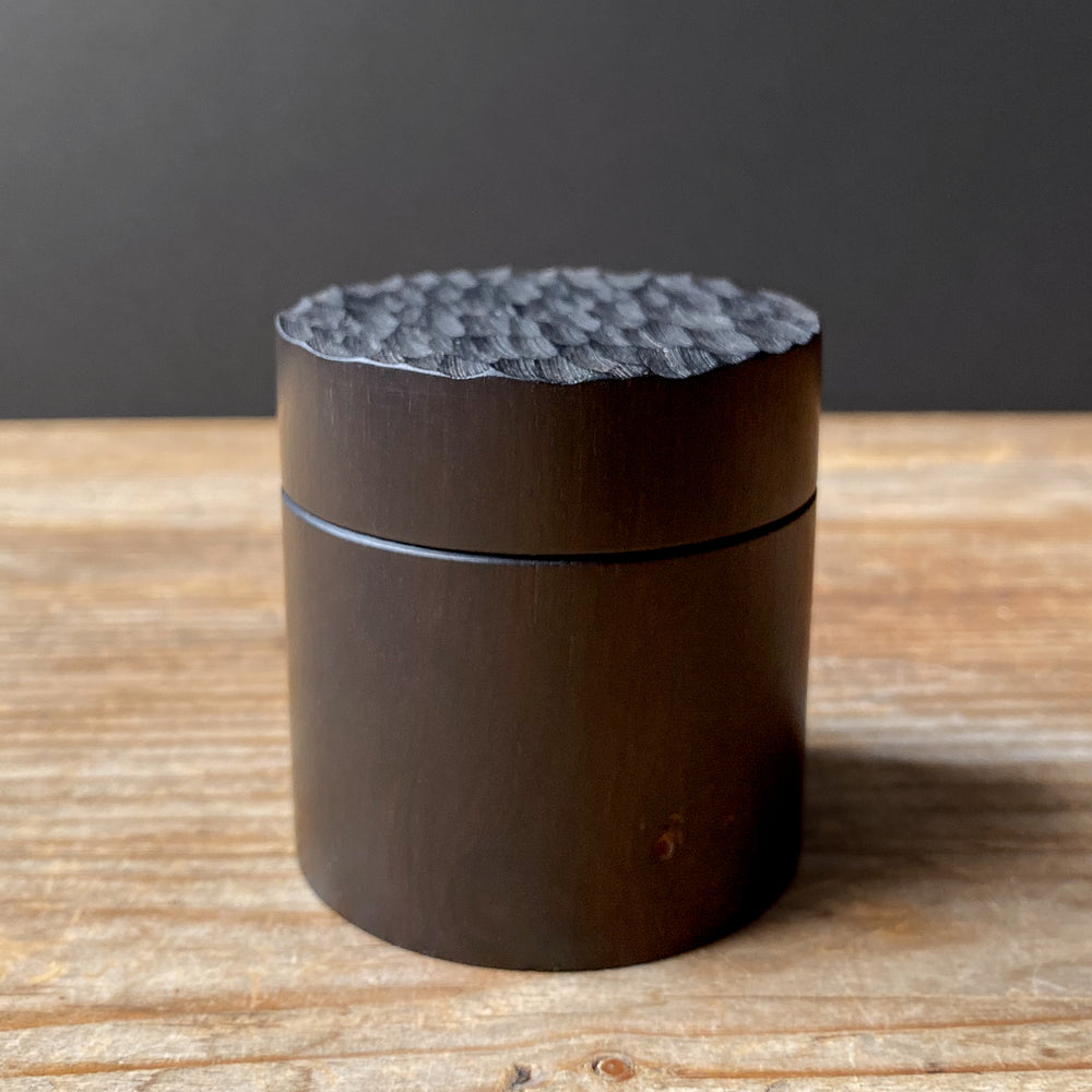 Small African Blackwood box with textured lid on wooden table against dark background.