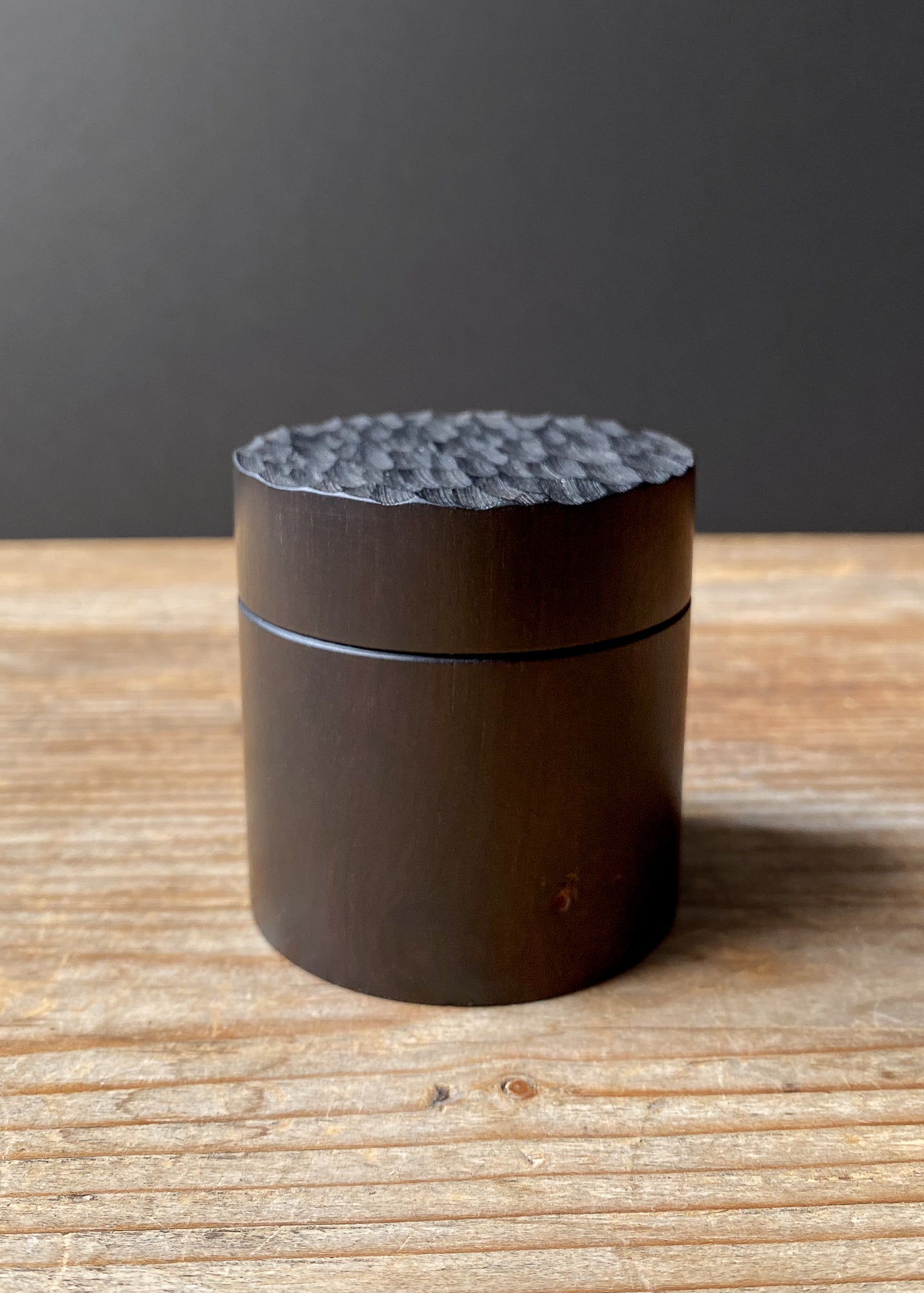 Small African Blackwood box with textured lid on wooden table against dark background.