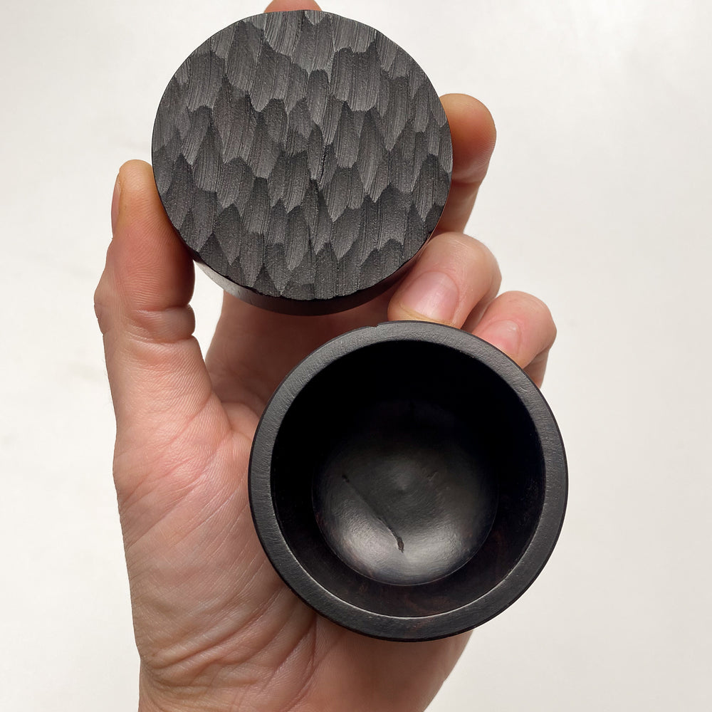 Hand holding opened small African Blackwood box with textured lid against white background.