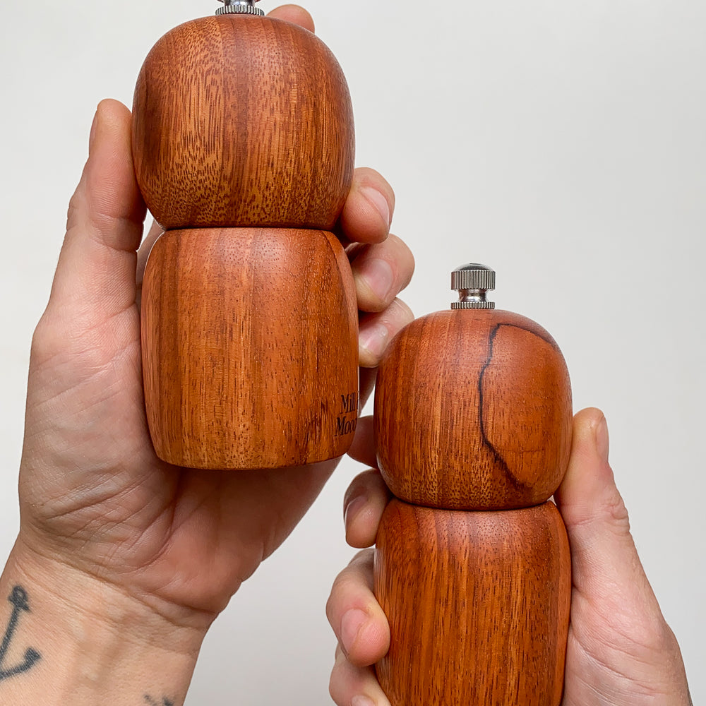 Two small reddish brown wooden pepper grinders held in hands against a plain background