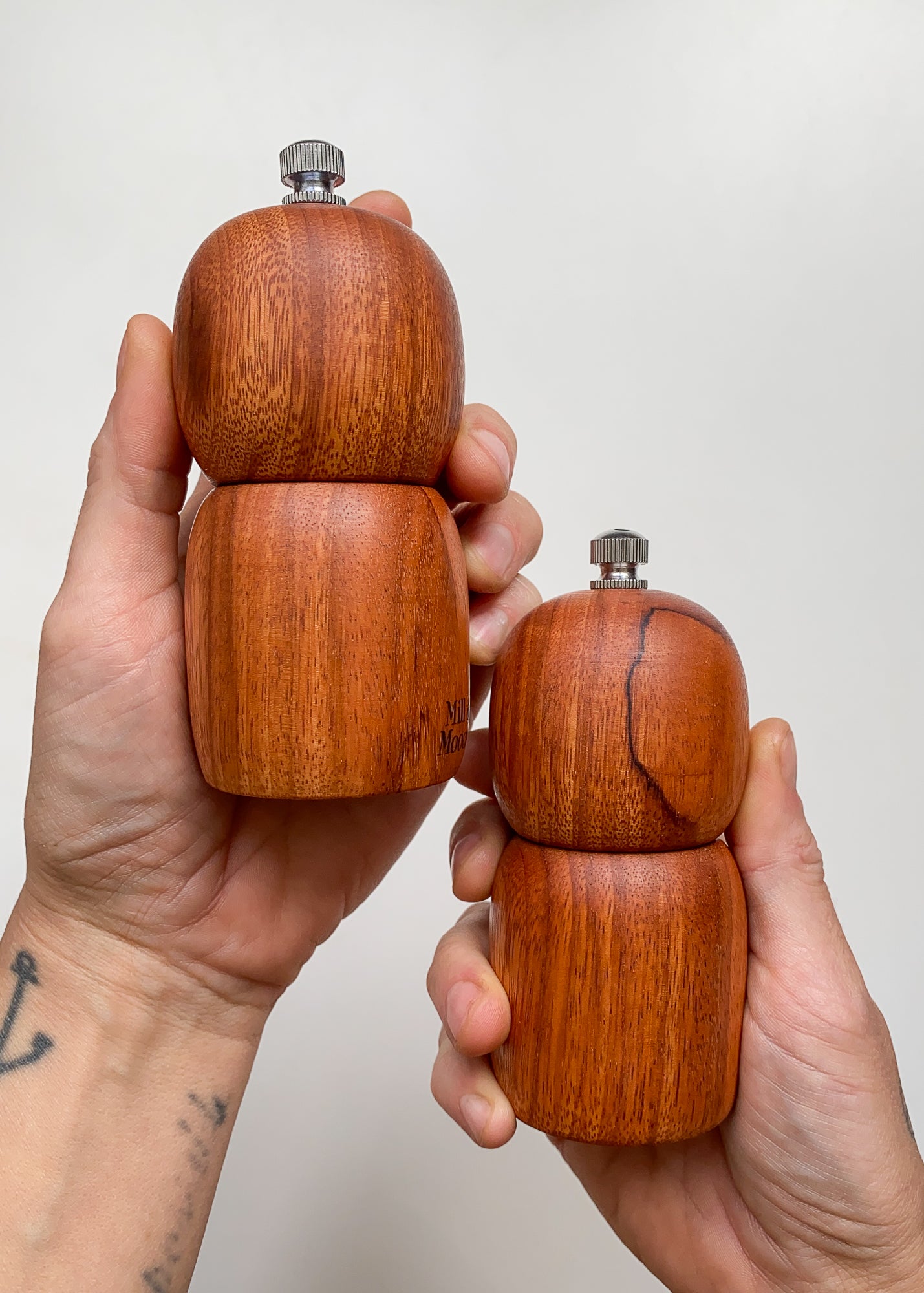 Two small reddish brown wooden pepper grinders held in hands against a plain background