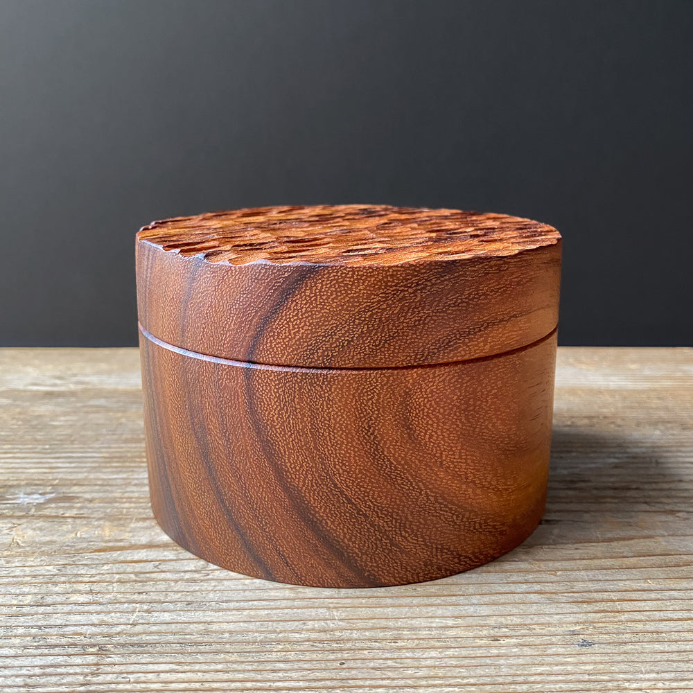 Cylindrical wooden box with textured lid on wood surface against dark background.