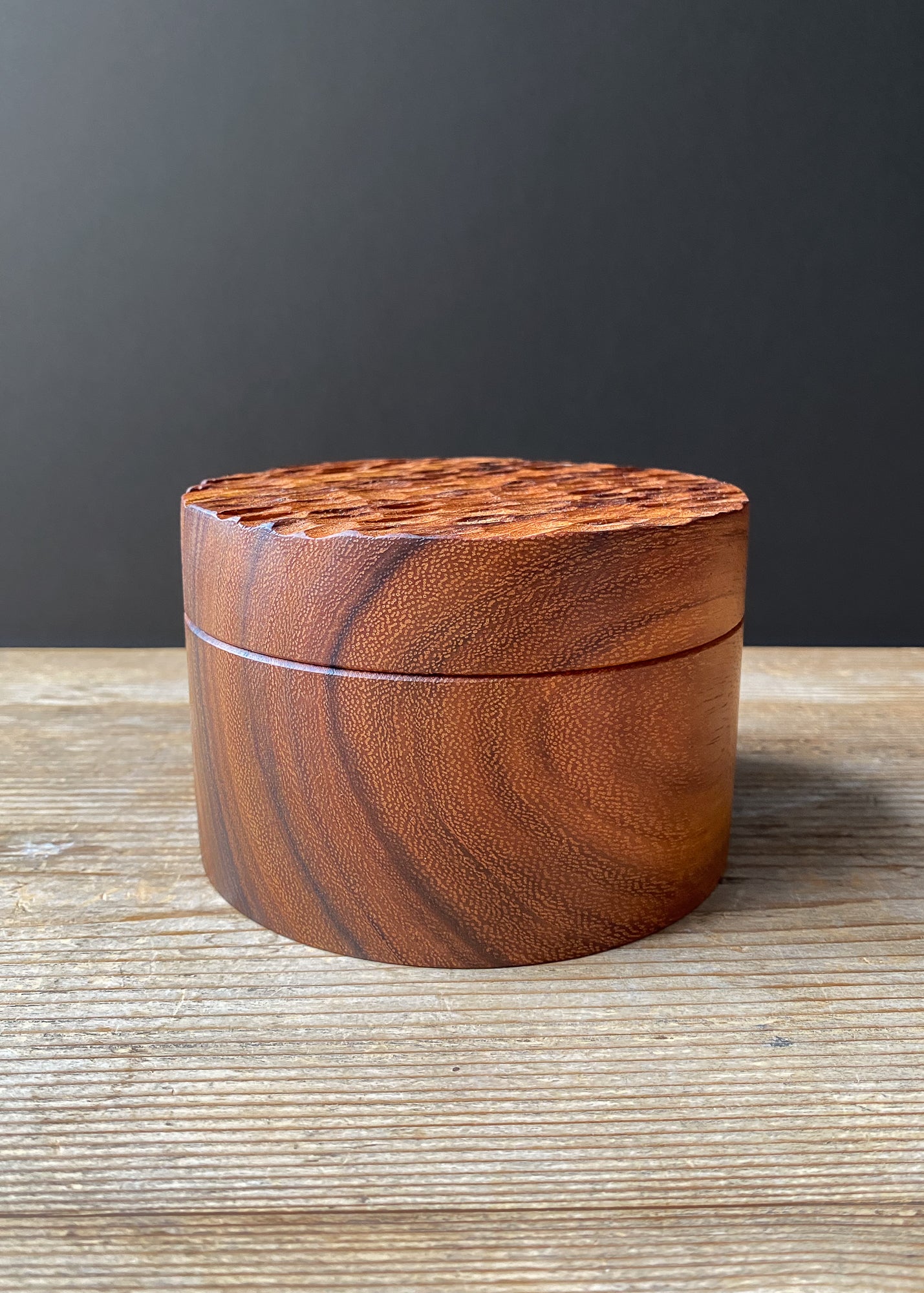 Cylindrical wooden box with textured lid on wood surface against dark background.