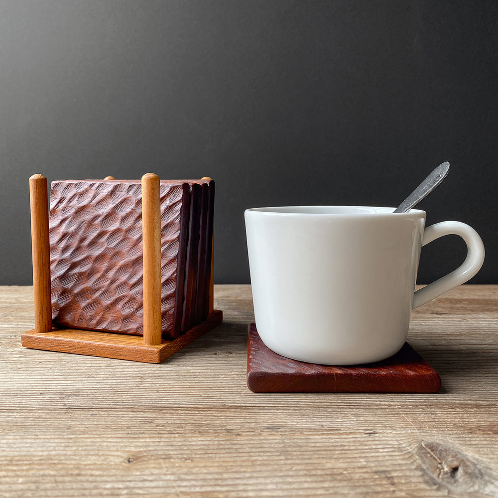 White mug with a spoon on a wooden coaster, next to a wooden coaster holder with textured coasters on a wooden surface.