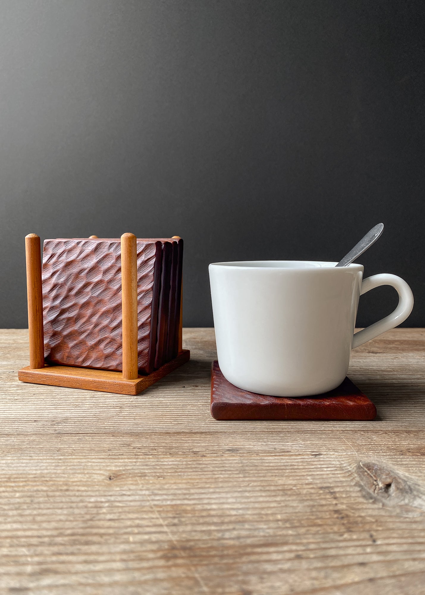 White mug with a spoon on a wooden coaster, next to a wooden coaster holder with textured coasters on a wooden surface.