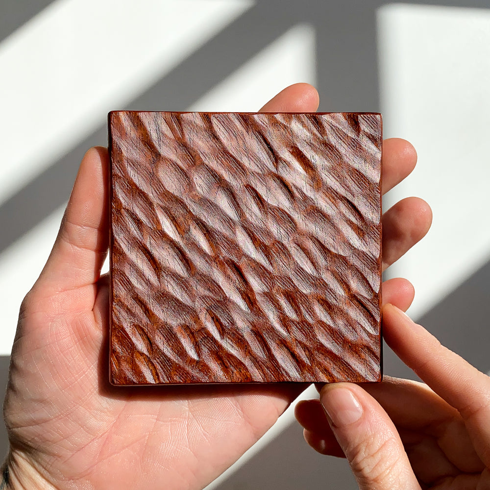 Hand holding a square wooden coaster with textured pattern against a neutral background