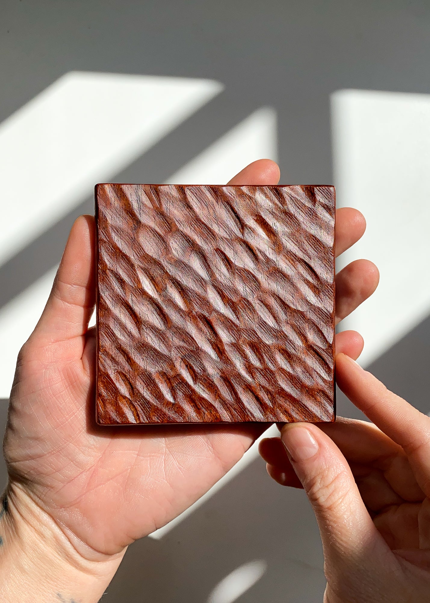 Hand holding a square wooden coaster with textured pattern against a neutral background
