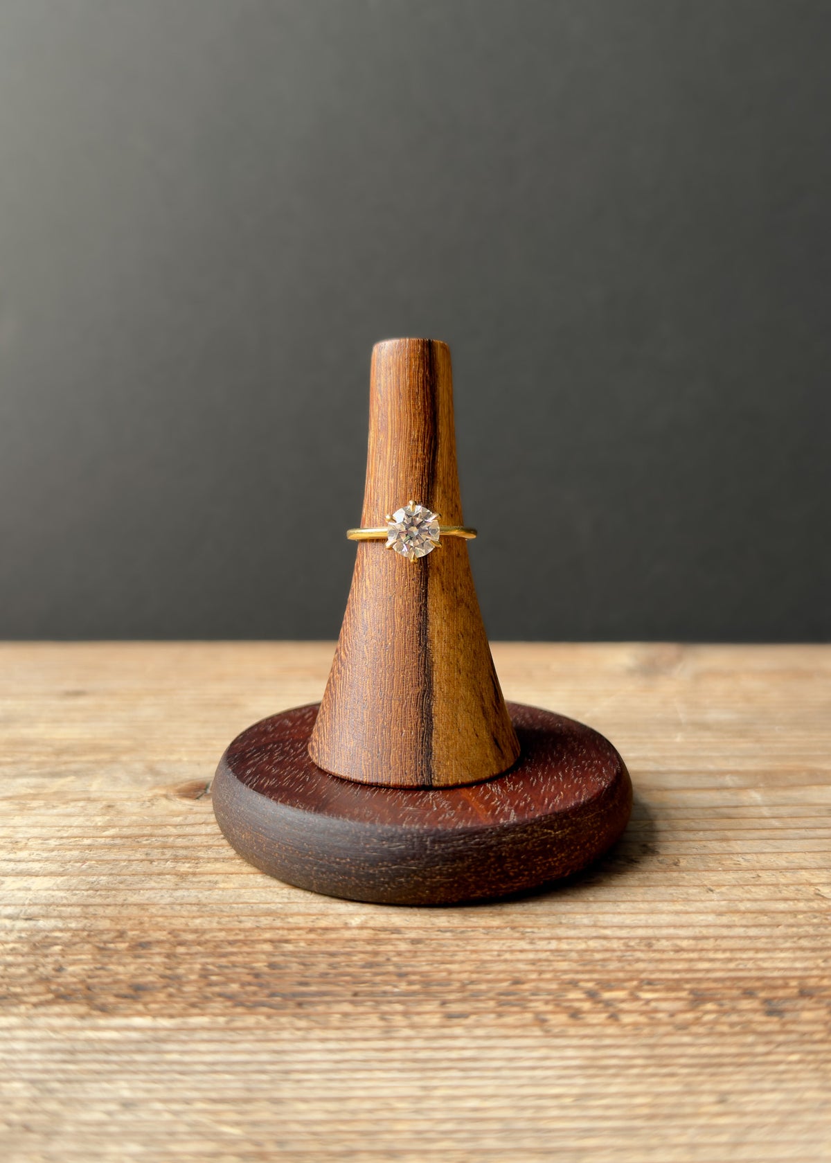 Two-tone wooden ring stand with conical top holding diamond ring on dark round wooden base against rustic background.