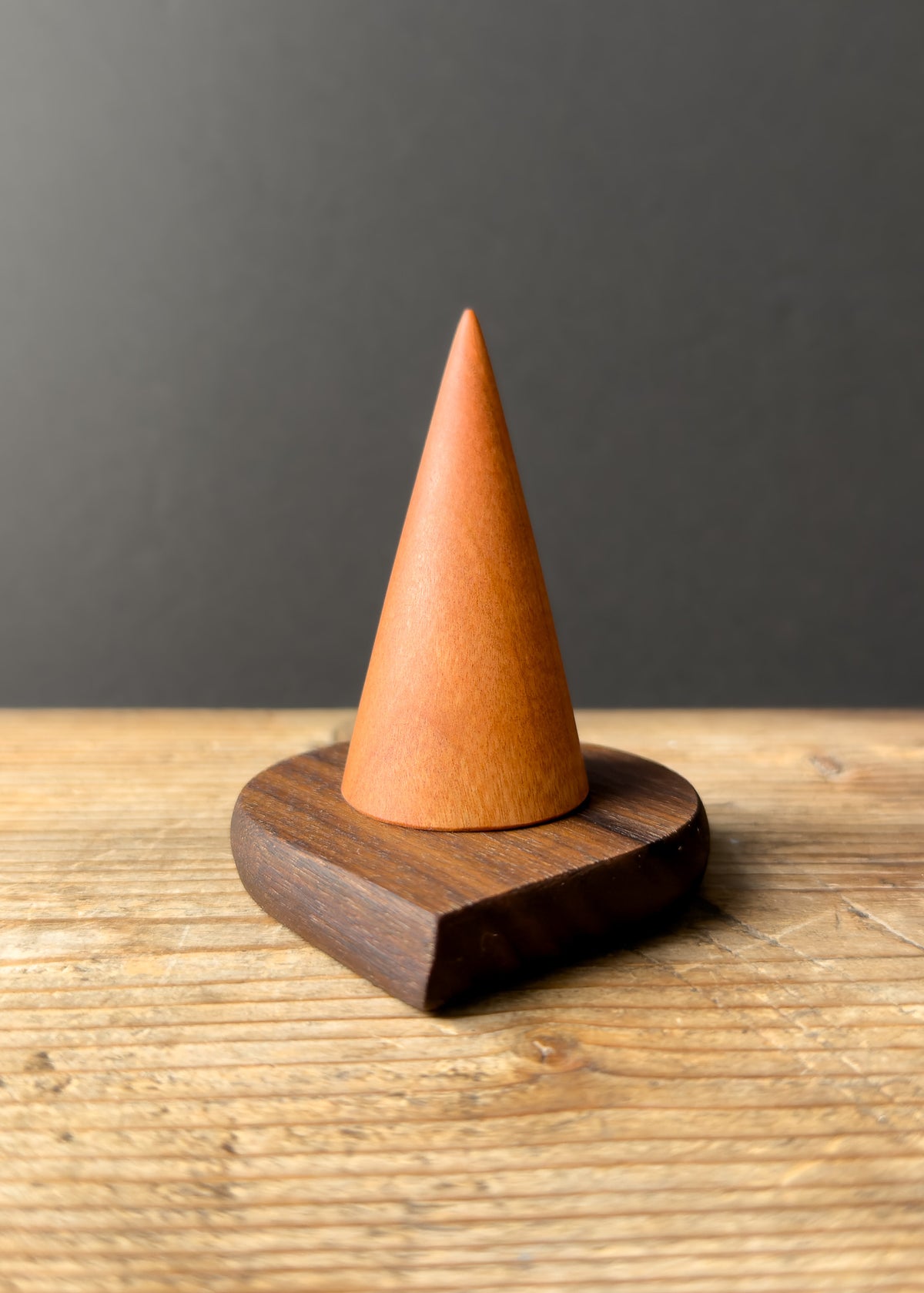 Wooden ring stand with pink pointed conical top and teardrop-shaped base on wooden table against dark background.
