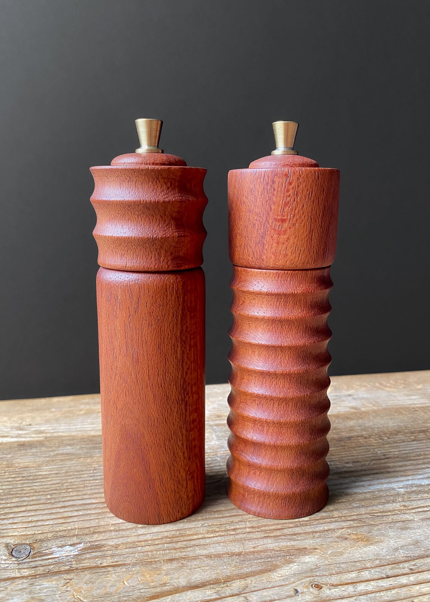 Two reddish brown wooden pepper mills on a wooden surface with a dark background