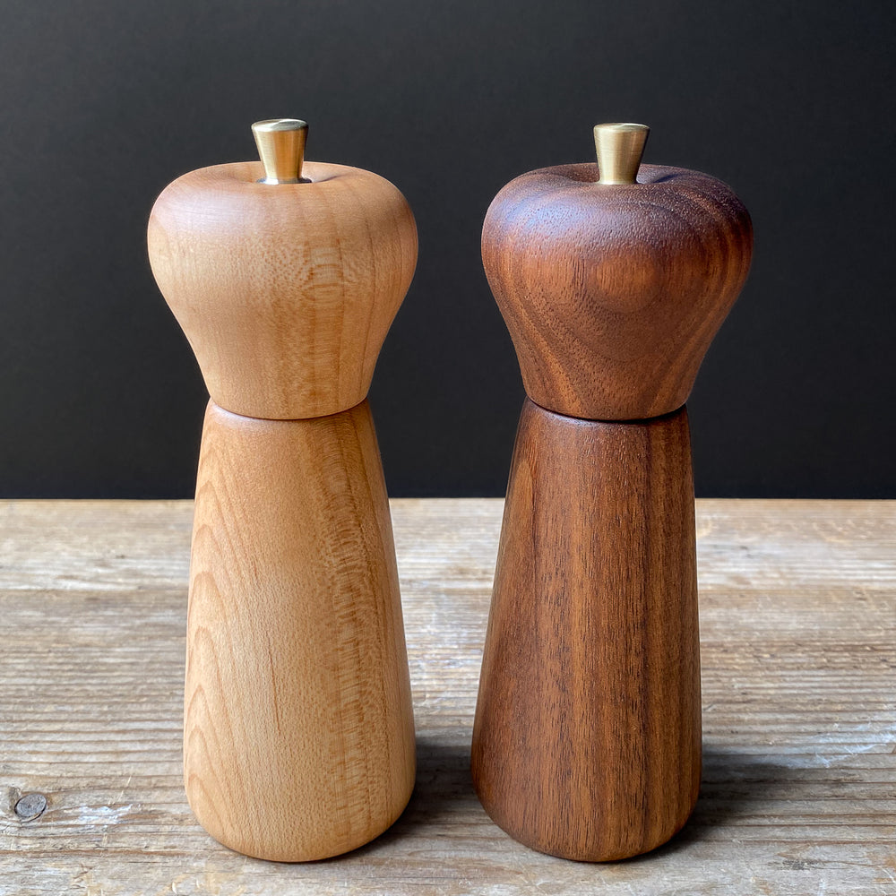 Two wooden pepper mills, one light brown and one dark brown, on a wooden surface with a dark background.