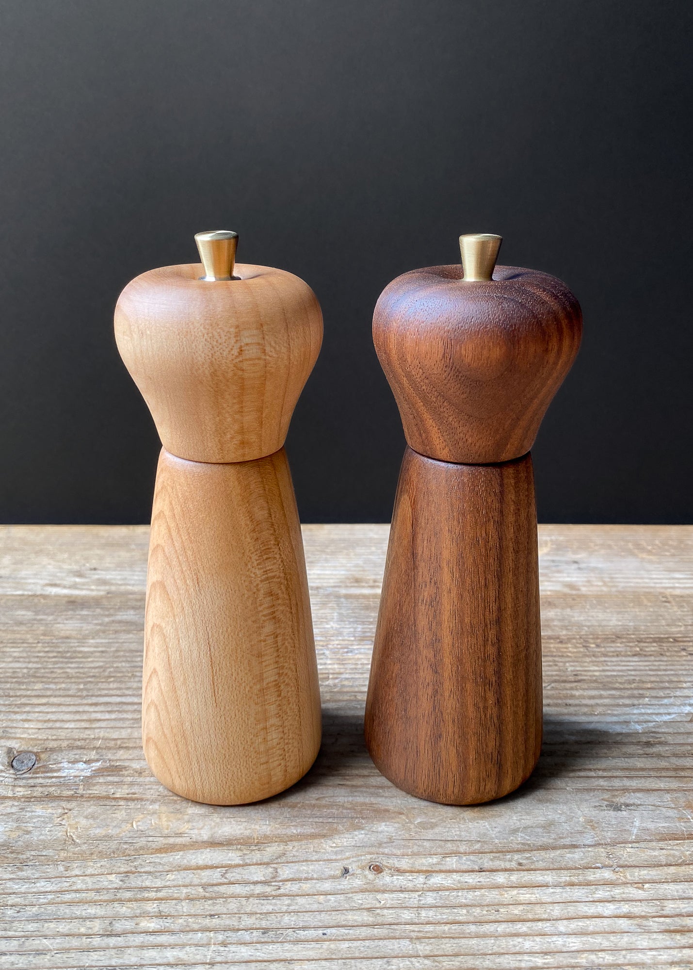 Two wooden pepper mills, one light brown and one dark brown, on a wooden surface with a dark background.