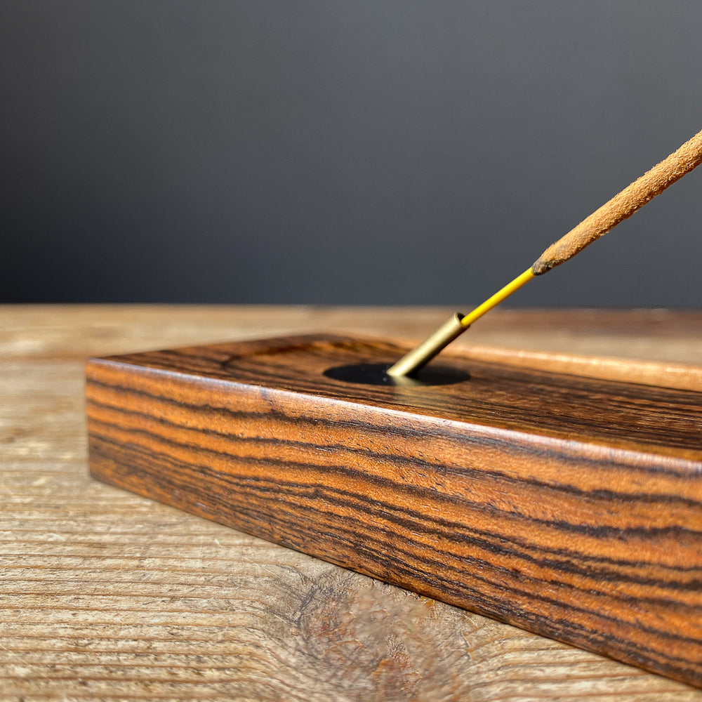 Close up side view of Bocote wood incense holder with brass detail and incense stick on wood table.