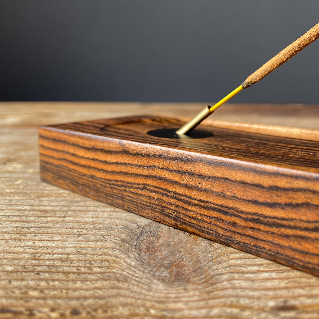 Close up view of Bocote incense holder with brass detail and long incense stick on wooden table. 